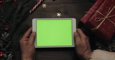 Claus Santa yeşil ekran beyaz tablet bilgisayarınızla tutan eller. Bazı mevcut kutuları masaya yerleştirerek diğer eller. Chroma anahtar. Üstten görünüm. Yılbaşı yeni yıl Internet sipariş kavramı