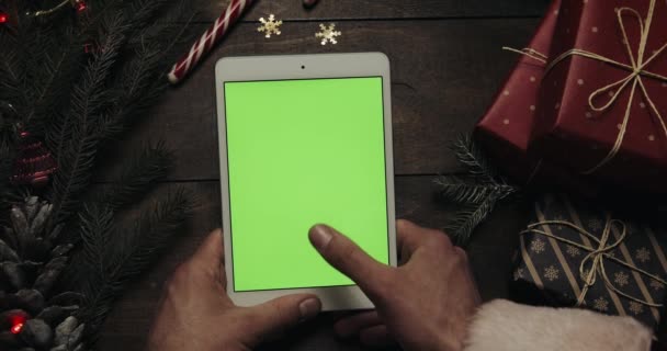 Руки Санта-Клауса держат белый планшетный компьютер с зеленым экраном. Другие руки кладут коробки на стол. Хрома-ключ. Вид сверху. Рождественский новогодний интернет-заказ — стоковое видео