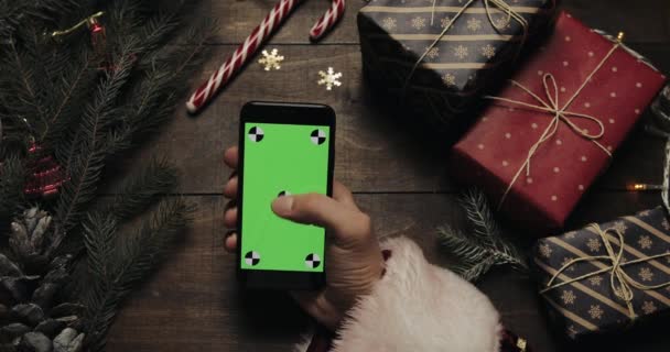 Santa Claus megérinti a fekete smartphone-val zöld képernyő oldalak kezében. Más kezek, néhány jelenlegi doboz elhelyezése az asztalon. Chroma-kulcs. Felülnézet. Karácsonyi-újévi internetes rendelés koncepció — Stock videók