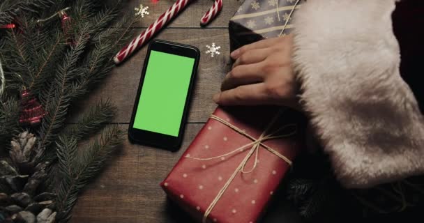 Vista superior. Smartphone preto com tela verde deitado na mesa com decoração de Natal feriado. Mãos de Papai Noel coloca um presente na mesa. Natal feriado decoração vista superior conceito — Vídeo de Stock