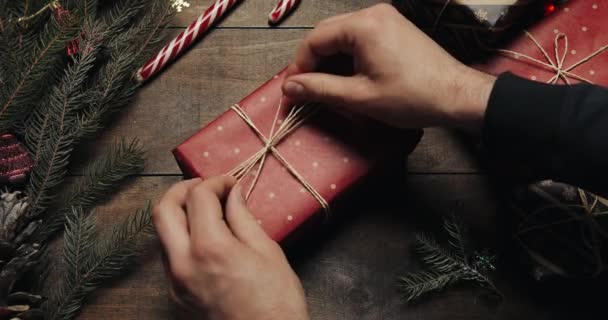 Vue du dessus des mains de l'homme attachant boîte cadeau avec fil de décoration après enveloppé de papier rouge sur la table en bois, se préparant à célébrer les vacances de Noël à la maison — Video