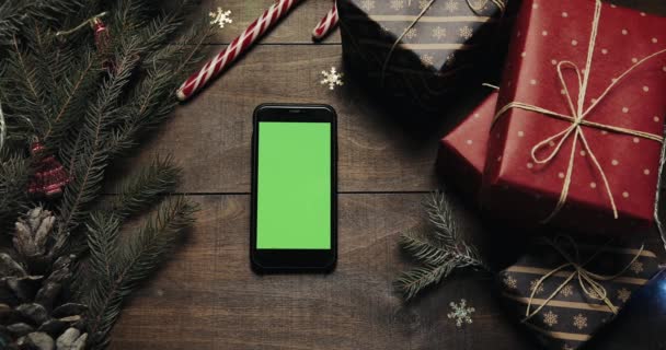 Topputsikt. Svart smarttelefon med grønn skjerm på bordet med julepynt. Julepynt, topp utsikt. Bakgrunn av tre – stockvideo
