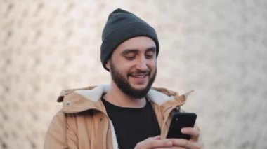 Shopping mall yakınındaki sokakta yürürken smartphone kullanan mutlu genç sakallı adam. O bir sonbahar ceket ve örme şapka giyiyor. İletişim, online alışveriş, sohbet, sosyal ağ kavramı.