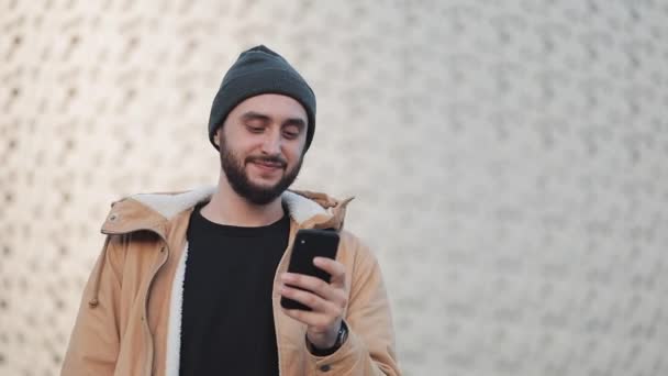 Feliz joven barba hombre usando teléfono inteligente caminando en la calle cerca del centro comercial. Lleva una chaqueta de otoño y un sombrero de punto. Comunicación, compras en línea, chat, concepto de redes sociales — Vídeos de Stock