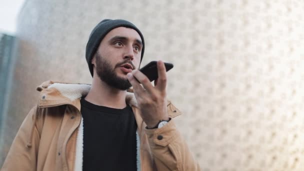 Giovane barbuto che invia messaggi audio sul cellulare all'aperto. Indossa una giacca autunnale e un cappello a maglia. Comunicazione, shopping online, chat, social network — Video Stock