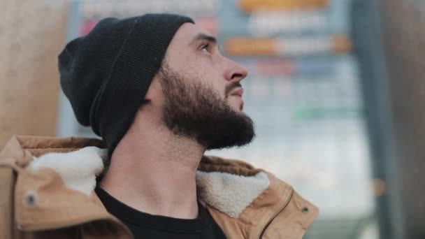Homem barba jovem passeio turístico na cidade viajando na Europa standinh perto de shopping center olha para a cidade. Ele está usando uma jaqueta de outono e chapéu de malha. Fechar — Vídeo de Stock