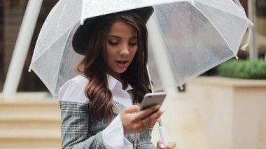 Yağışlı hava, gülümseyen, holding şemsiye, iletişim kavramı sokakta Smartphone kullanarak güzel genç iş kadını