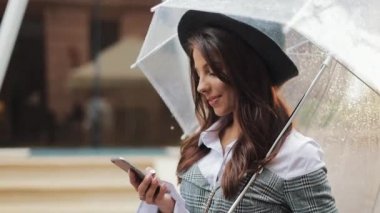 Gülümseyerek, şemsiye tutan yağmurlu havalarda sokakta smartphone kullanarak güzel genç iş kadını. Kameraya bakıyor. İletişim kavramı