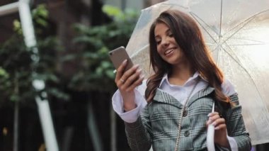 Yağışlı hava, gülümseyen, holding şemsiye, iletişim kavramı sokakta Smartphone kullanarak güzel genç iş kadını.