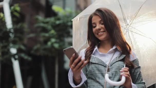 Молодая бизнесвумен, пользующаяся смартфоном на улице в дождливую погоду, улыбающаяся, держащая в руках пуховик, коммуникационная концепция — стоковое видео