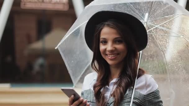 Schöne junge Geschäftsfrau mit Smartphone auf der Straße bei regnerischem Wetter, lächelnd, Regenschirm in der Hand. sie blickt in die Kamera. Kommunikationskonzept — Stockvideo