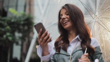 Yağışlı hava, gülümseyen, holding şemsiye, iletişim kavramı sokakta Smartphone kullanarak güzel genç iş kadını