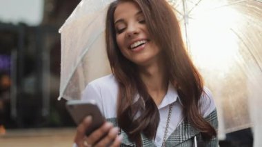 Yağışlı hava, gülümseyen, holding şemsiye, iletişim kavramı sokakta Smartphone kullanarak güzel genç iş kadını