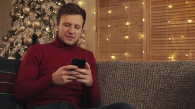 Mesajlaşma, yakın güzel bir şekilde dekore edilmiş daire içinde gülümseyen koltukta oturan smartphone kullanarak çekici bir adam. Noel ağacı