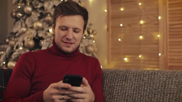 Bărbat atractiv folosind smartphone-ul stând pe canapea, mesagerie, zâmbind într-un apartament decorat lângă frumos. Pomul de Crăciun — Videoclip de stoc