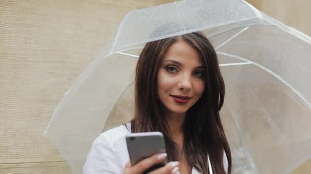 Красива молода бізнес-леді, використовуючи смартфон на вулиці в дощову погоду, посміхається, тримає парасольку. Вона дивиться в камеру. Концепція комунікації — стокове відео