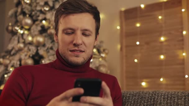 Attraktiver Mann mit Smartphone auf der Couch sitzend, Nachrichten, lächelnd in der geschmückten Wohnung in der Nähe nett. Weihnachtsbaum — Stockvideo