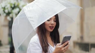 Gülümseyerek, şemsiye tutan yağmurlu havalarda sokakta yürürken smartphone kullanarak güzel genç iş kadını. İletişim kavramı