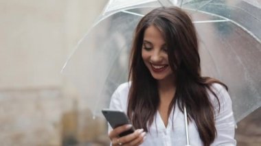 Gülümseyerek, şemsiye tutan yağmurlu havalarda sokakta yürürken smartphone kullanarak güzel genç iş kadını. İletişim kavramı