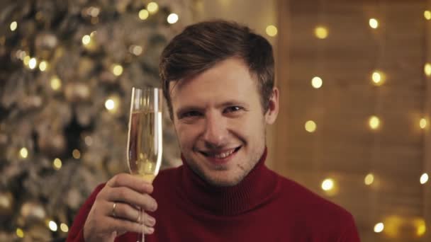 Atractivo hombre con una copa de champán mirando a la cámara en el fondo del árbol de Navidad. Levanta la copa. Movimiento lento — Vídeos de Stock