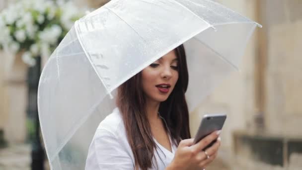 Красива молода бізнес-леді, використовуючи смартфон, що йде по вулиці в дощову погоду, посміхається, тримає парасольку. Концепція комунікації — стокове відео