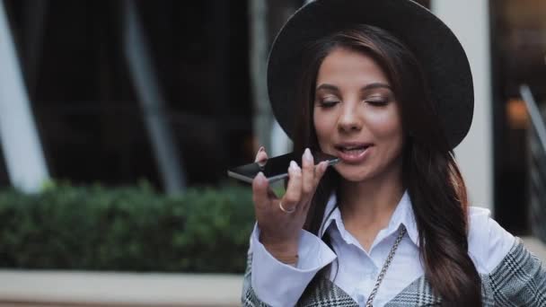 Femme d'affaires en chapeau noir envoyer un message vocal audio sur le téléphone portable à l'extérieur parler à l'assistant mobile. Fille en utilisant la reconnaissance vocale smartphone, dicte pensées, message de composition vocale — Video