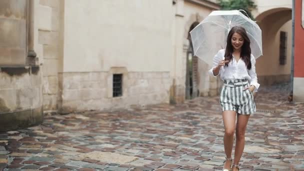 Улыбающаяся юная брюнетка в платье ходит с зонтиком по улице старого города. Прогулка под дождем — стоковое видео