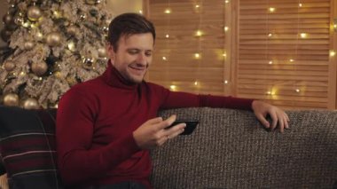Mesajlaşma, yakın güzel bir şekilde dekore edilmiş daire içinde gülümseyen koltukta oturan smartphone kullanarak çekici bir adam. Aygıt video izlerken. Noel ağacı arka plan