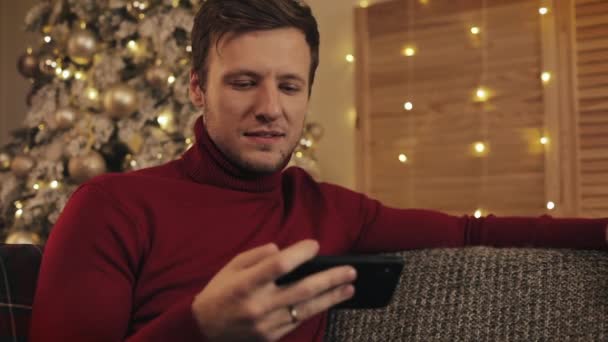 Hombre atractivo usando teléfono inteligente sentado en el sofá, mensajería, sonriendo en el apartamento decorado cerca de agradable. Está viendo el vídeo en el dispositivo. Árbol de Navidad fondo — Vídeos de Stock