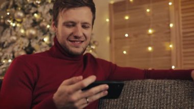 Mesajlaşma, yakın güzel bir şekilde dekore edilmiş daire içinde gülümseyen koltukta oturan smartphone kullanarak çekici bir adam. Aygıt video izlerken. Noel ağacı arka plan