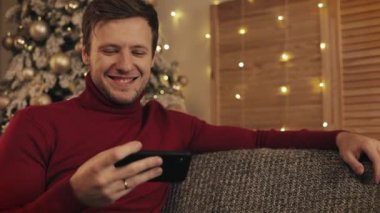 Mesajlaşma, yakın güzel bir şekilde dekore edilmiş daire içinde gülümseyen koltukta oturan smartphone kullanarak çekici bir adam. Aygıt video izlerken. Noel ağacı arka plan