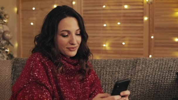 Mujer atractiva usando teléfono inteligente sentado en el sofá, mensajería, sonriendo en el apartamento decorado cerca de agradable. Árbol de Navidad fondo — Vídeo de stock