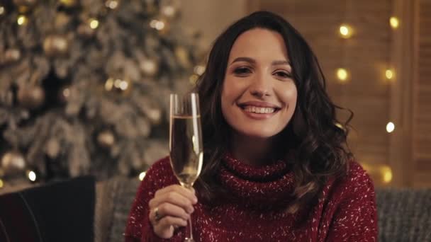 Attraktive Frau mit einem Glas Champagner, die in die Kamera auf dem Hintergrund des Weihnachtsbaums blickt. hebt sie das Glas. Zeitlupe — Stockvideo