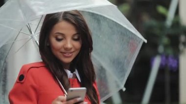 Yağışlı hava, gülümseyen, holding şemsiye, iletişim kavramı sokakta Smartphone kullanarak güzel genç iş kadını