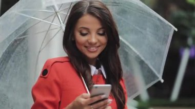 Yağışlı hava, gülümseyen, holding şemsiye, iletişim kavramı sokakta Smartphone kullanarak güzel genç iş kadını