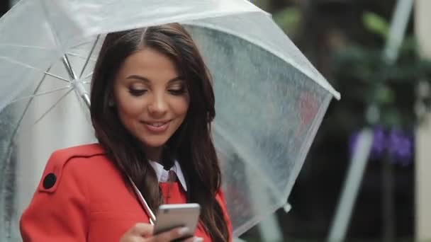 Hermosa joven mujer de negocios utilizando el teléfono inteligente en la calle en tiempo lluvioso, sonriendo, sosteniendo el paraguas, concepto de comunicación — Vídeo de stock