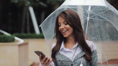 Gülümseyerek, şemsiye tutan yağmurlu havalarda sokakta yürürken smartphone kullanarak mutlu genç iş kadını. İletişim kavramı