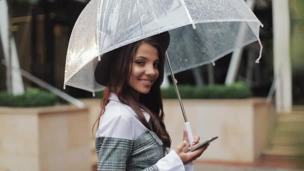 专业的年轻商务女士站在城市街道上, 使用智能手机。她看着镜头, 拿着伞。理念: 新业务、沟通、银行家。外面, 慢动作 — 图库视频影像