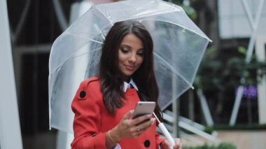 Kentsel Caddesi üzerinde duran ve kırmızı palto giyiyor smartphone kullanarak mutlu genç iş kadını. O holding şemsiyesi onun elinde. Konsept: yeni iş, iletişim, bankacı. Dış, ağır çekim