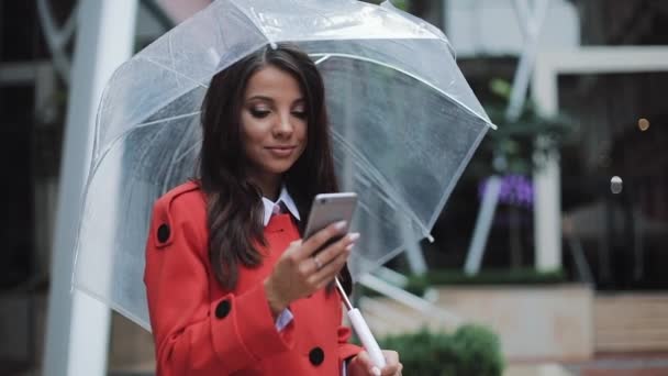 Feliz joven mujer de negocios de pie en la calle urbana y el uso de teléfonos inteligentes con abrigo rojo. Ella sosteniendo el paraguas en sus manos. Concepto: nuevo negocio, comunicación, banquero. Afuera, cámara lenta — Vídeo de stock