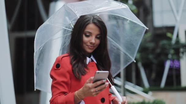 Щаслива молода бізнес-леді, що стоїть на міській вулиці і використовує смартфон у червоному пальто. Вона тримає парасольку в руках. Концепція: новий бізнес, комунікація, банкір. Зовні, повільний рух — стокове відео