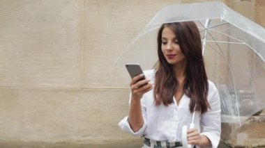 Yağışlı hava, gülümseyen, holding şemsiye, iletişim kavramı sokakta Smartphone kullanarak güzel genç iş kadını