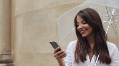 Yağışlı hava, gülümseyen, holding şemsiye, iletişim kavramı sokakta Smartphone kullanarak güzel genç iş kadını