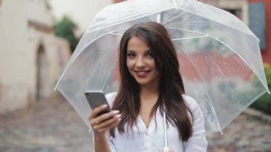 Gülümseyerek, şemsiye tutan yağmurlu havalarda sokakta smartphone kullanarak güzel genç iş kadını. Kameraya bakıyor. İletişim kavramı