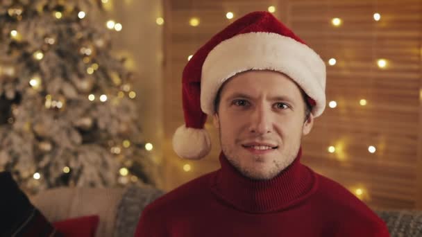 Портрет улыбающегося человека в Санта-Клаусе, смотрящего в камеру на фоне рождественских деревьев. Медленное движение — стоковое видео
