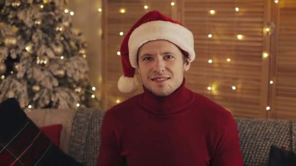 Retrato del hombre atractivo en Santa Sombrero Mirando a la cámara en el fondo del árbol de Navidad. Movimiento lento — Vídeos de Stock