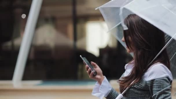雨の天候、笑みを浮かべて、持株傘、コミュニケーション コンセプトの通りを歩いてのスマート フォンを使用して美しい若いビジネス女性 — ストック動画