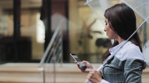 Piękny, młody biznes kobieta przy użyciu smartfona spaceru na ulicy w deszczowa pogoda, uśmiechnięty, parasol gospodarstwa, komunikacja koncepcja — Wideo stockowe