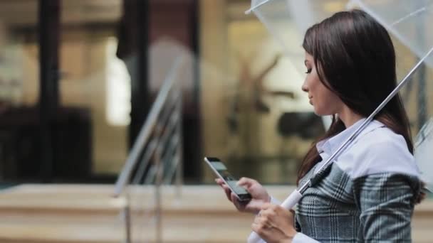 Hermosa joven mujer de negocios usando teléfono inteligente caminando por la calle en tiempo lluvioso, sonriendo, sosteniendo el paraguas, concepto de comunicación — Vídeos de Stock