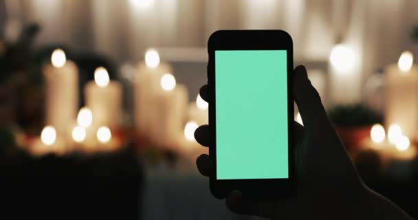 Male Hand is Holding Smartphone with Green Screen. Lighted candles in the background — Stock Video